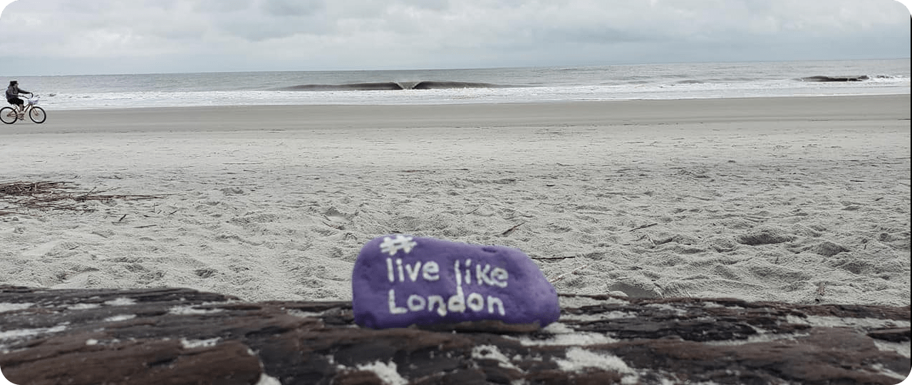 A purple Live Like London rock resting on rocks at the shoreline, with a cyclist riding past on the beach.