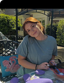 A smiling young painter at a backyard table with finished purple rocks and a flamingo painting.