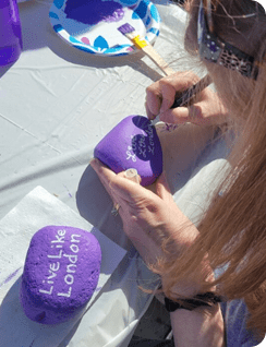 A young woman painting a Live Like London message on a purple rock outdoors.
